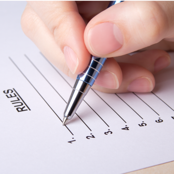 A hand holding a pen hovers over a sheet of paper titled “RULES,” which lists numbers 1 through 7 with blank lines beside each number, ready to be filled in.