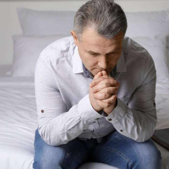 Person sitting on a bed, hands clasped, with a Bible beside them.