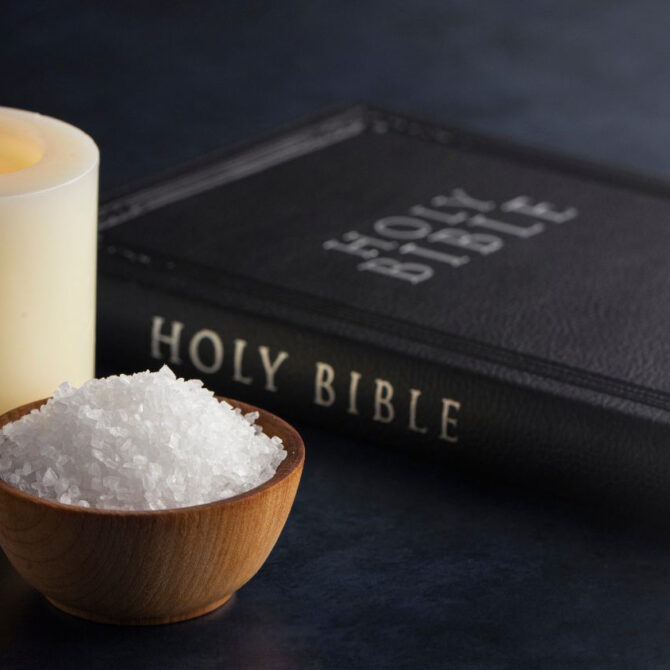 A lit white candle, a wooden bowl filled with coarse white salt, and a black leather Holy Bible arranged on a dark surface, with the candle’s warm light illuminating the scene.