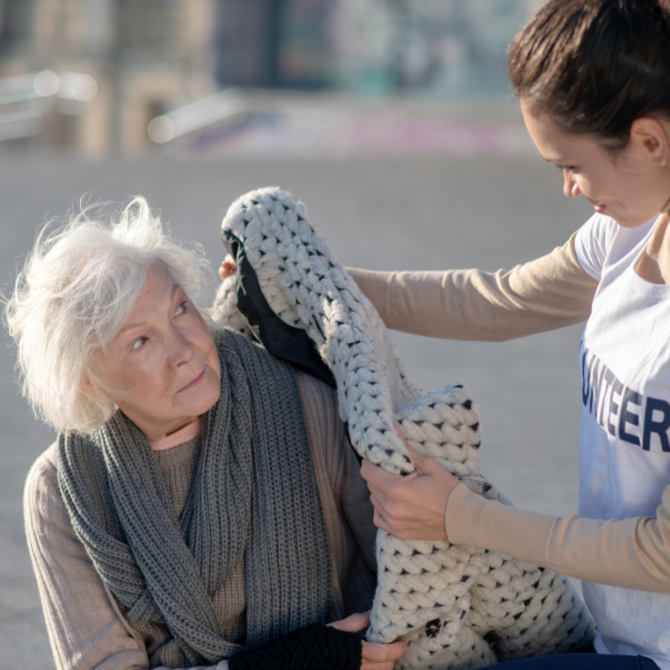 A volunteer wearing a white shirt labeled “VOLUNTEER” hands a thick knitted blanket to an older person with gray hair who is sitting outdoors. The older person is dressed in a warm sweater and scarf. They appear to be in an urban setting with pavement and blurred buildings in the background. The scene conveys kindness and assistance.