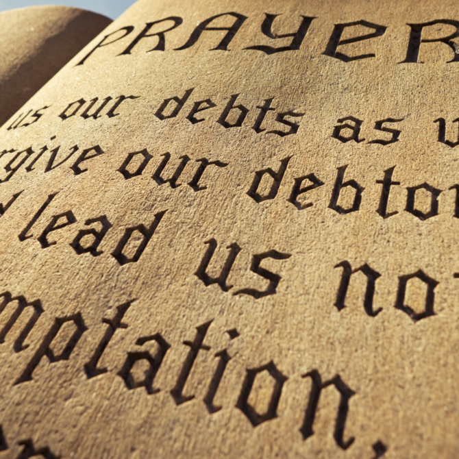 Close-up of a stone tablet engraved with large black-letter text, including the word “PRAYER” near the top. The weathered, sand-colored surface is shown at an angle, with parts of the inscription in focus and other areas blurred by shallow depth of field. Sunlight highlights the textured stone against a clear blue sky in the background.