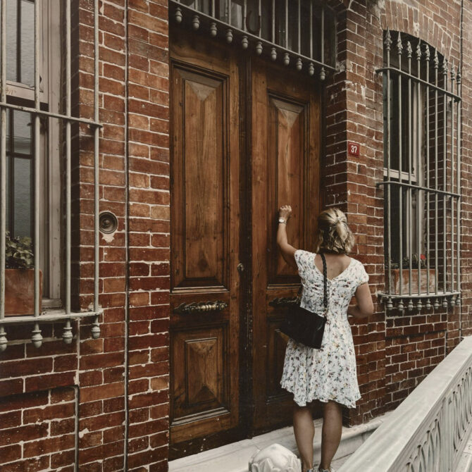 A person stands on a stone walkway and knocks on large wooden double doors of a historic red brick building.